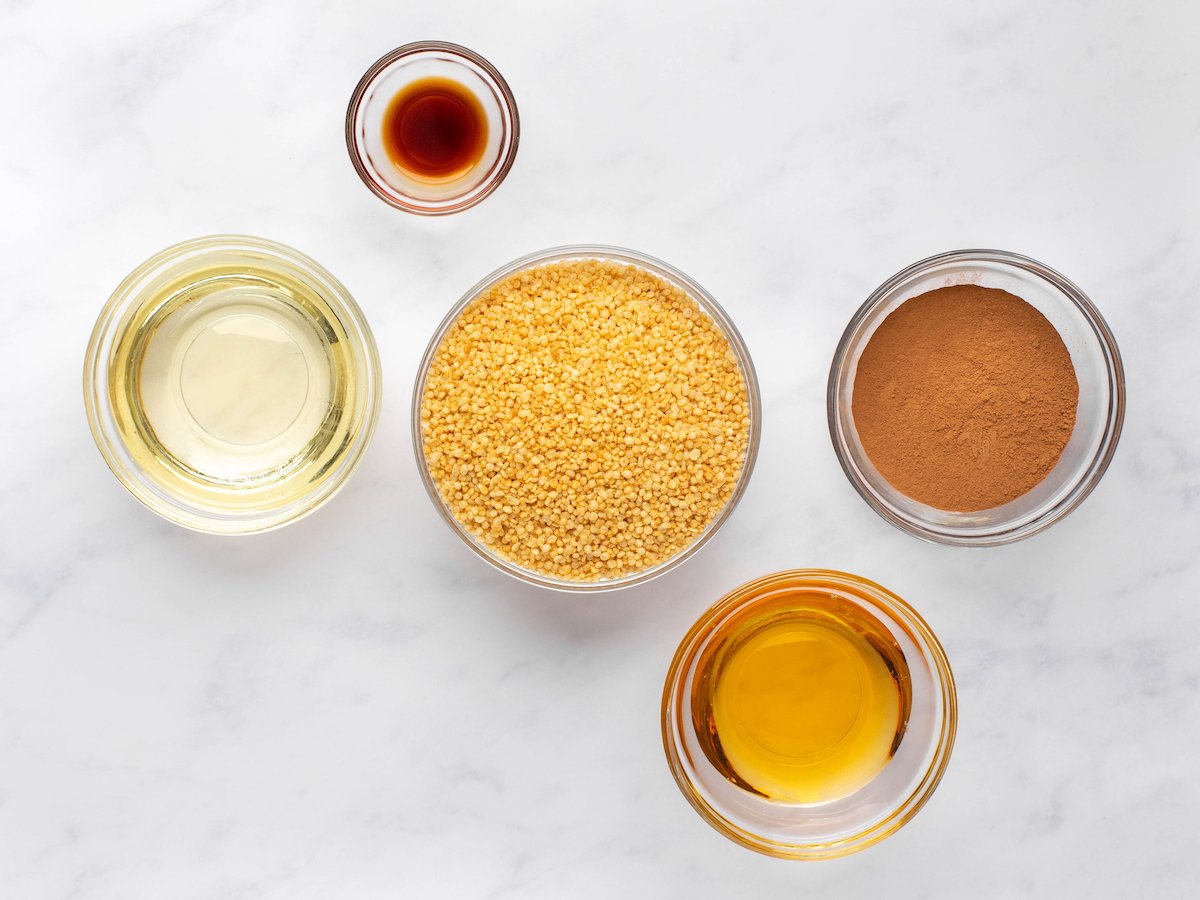 This is an overhead image of five glass bowls on a white marble surface. The small bowls have vanilla, melted coconut oil, quinoa puffs, cocoa powder, and agave nectar.