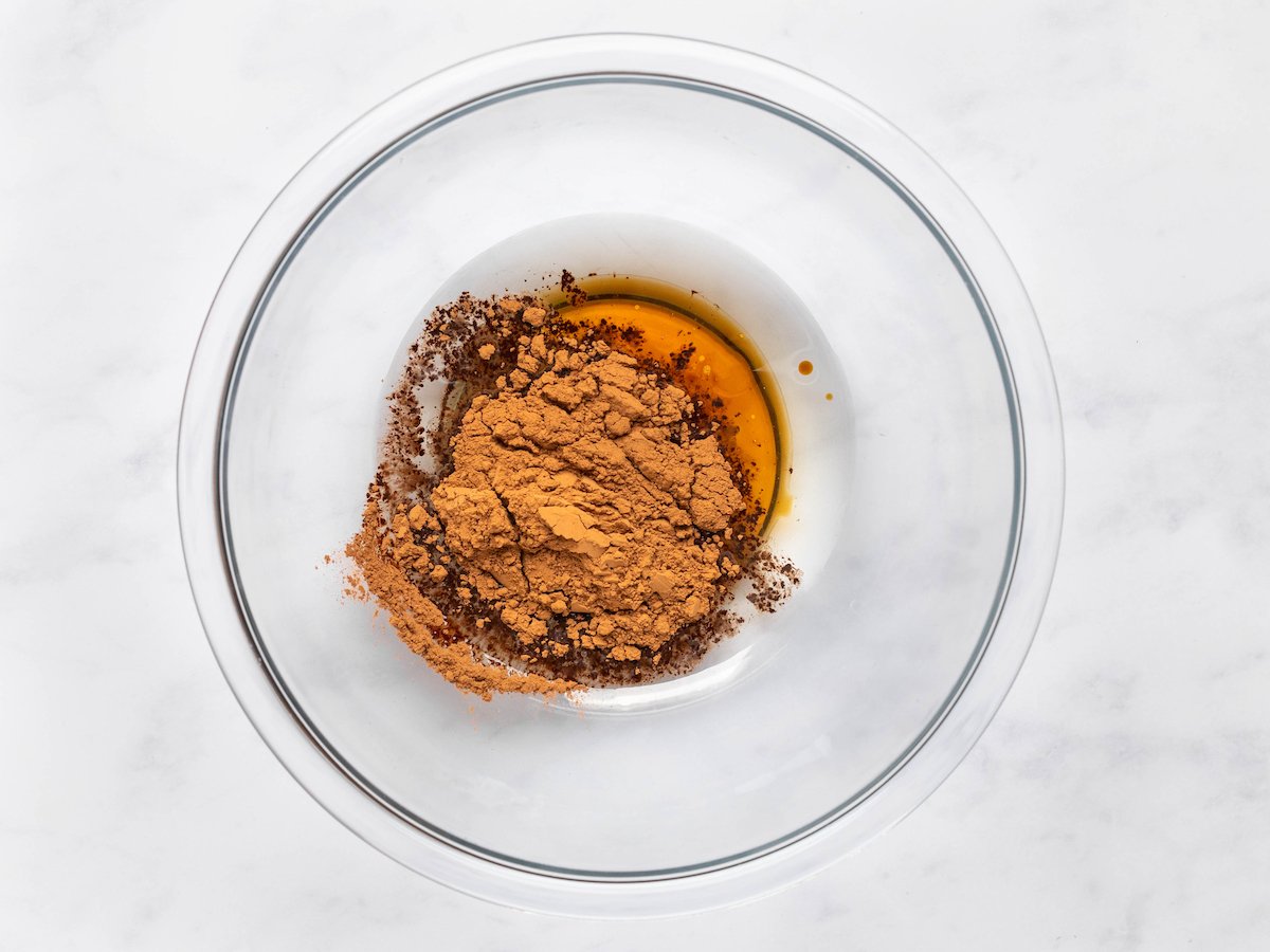 This is a horizontal overhead image of a glass bowl with melted coconut oil, agave, and cocoa powder. The glass bowl sits on a white marble surface.