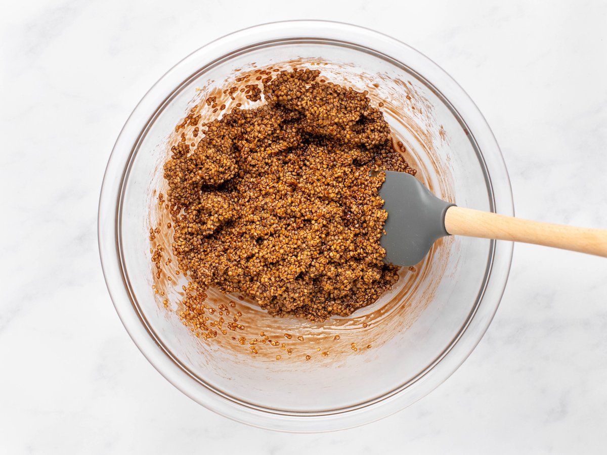 This is an overhead horizontal image of a glass bowl filled with a chocolate and quinoa mixture and a wood and grey rubber spatula leaning against the side of the bowl. The glass bowl sits on a white marble counter.