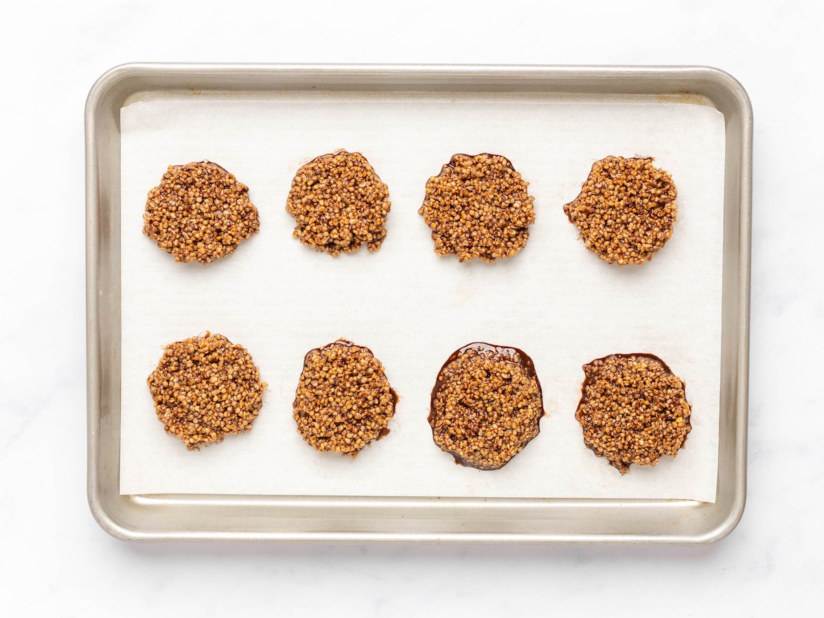This is a horizontal image of chocolate quinoa circles on a baking sheet with parchment paper. The chocolate around the quinoa is melty. The baking sheet sits on a white marble surface.
