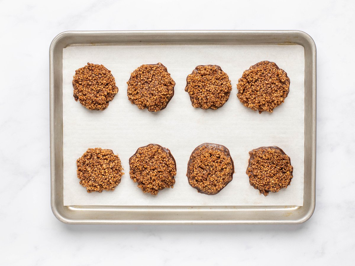 This is a horizontal image of chocolate quinoa circles on a baking sheet with parchment paper. The chocolate surrounding the quinoa is hardened. The baking sheet sits on a white marble surface.