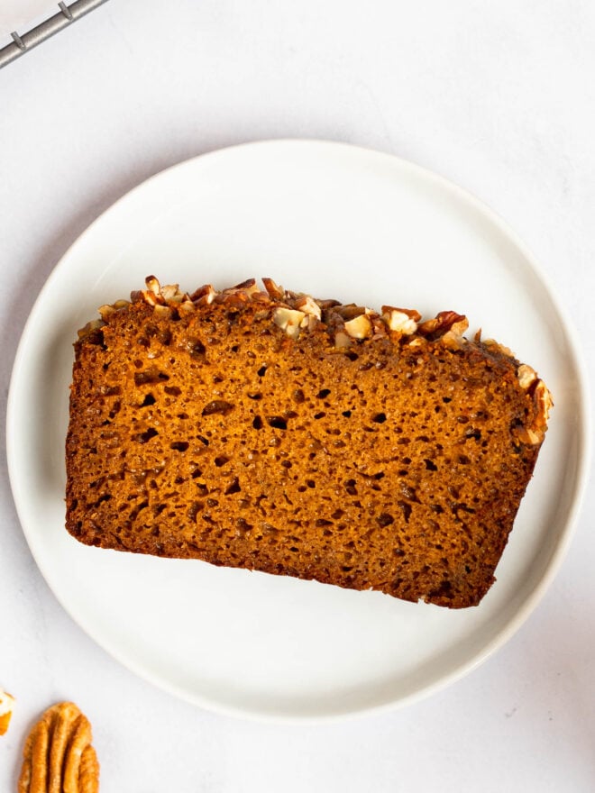 This is an overhead image of a white plate with a slice of sweet potato bread on it. You can see the light, fluffy texture of the bread with bubbles. You can also see the top crust layer of the bread that is topped with crushed pecans. The plate sits on a white surface with the edge of a cooling rack to the top left corner of the image and a single pecan to the bottom left.