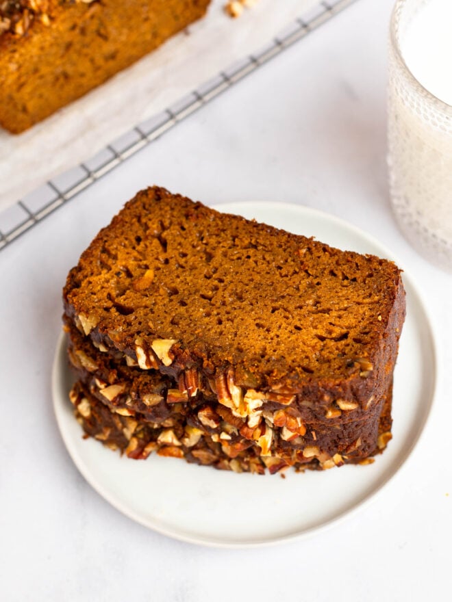 This is an angled 3/4 view vertical image of a white plate with slices of sweet potato bread stacked on it. The bread slices has chopped pecans on top. The plate sits on a white marble surface. A cooling rack is blurred in the background, to the top left corner of the image with white parchment paper and the rest of the bread loaf on top. To the right top corner of the image is a glass of milk.