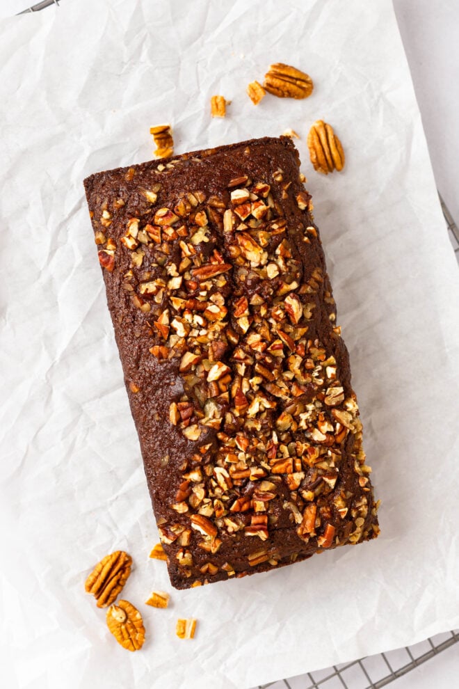 This is an overhead vertical image of a sweet potato bread topped with crushed pecans. The bread sits on a crumbled white piece of parchment paper on a silver cooling rack on a white marble surface. More pecans are around the bread, on the parchment paper.