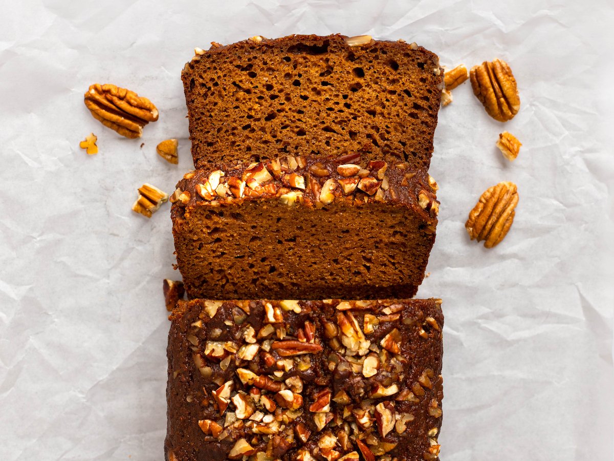 This is an overhead horizontal image of a sweet potato bread with chopped pecans on top. The bread is coming up from the bottom center of the image and has two slices cut and down on the crumbled piece of parchment paper under the bread. More pecans are on the parchment paper around the bread. 
