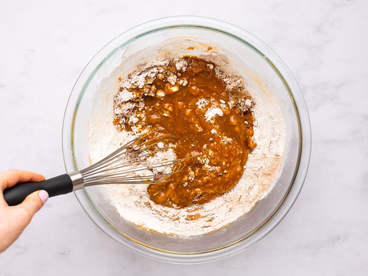 This is an overhead horizontal image of a glass bowl sitting on a white marble surface. In the bowl is a light brown/dark orange batter with dry ingredients being whisked into the mixture. A whisk is in the batter with the handle being held to the left side of the image.