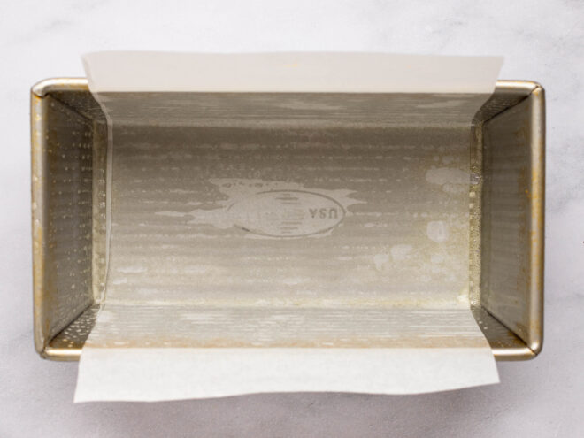 This is an overhead horizontal image of a bread pan with white parchment paper hanging over the two long sides of the pan. The bread pan sits horizontally on a white marble surface.