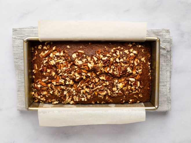 This is an overhead horizontal image of a bread pan with a light brown bread with crushed pecans on top. White parchment paper hangs over the long sides of the pan. The pan sits on a folded light grey striped tea towel on a white marble surface.