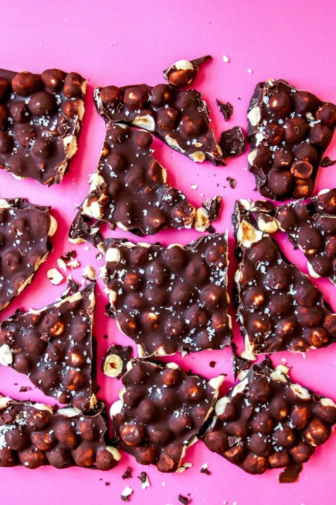 This is a vertical overhead image of chocolate hazelnut bark on a dark pink surface. The bark is broken into un-uniform pieces and sprinkled with flakey salt on top. 
