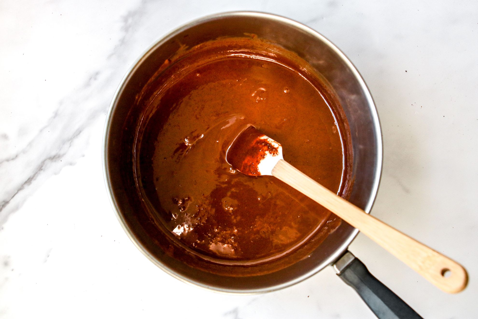 This is an overhead image of a saucepan with melted chocolate in it. The pot sits on a white marble surface. A small white rubber spatula is dipped into the chocolate and the wooden handle is leaning against the side of the pot, to the right bottom corner.