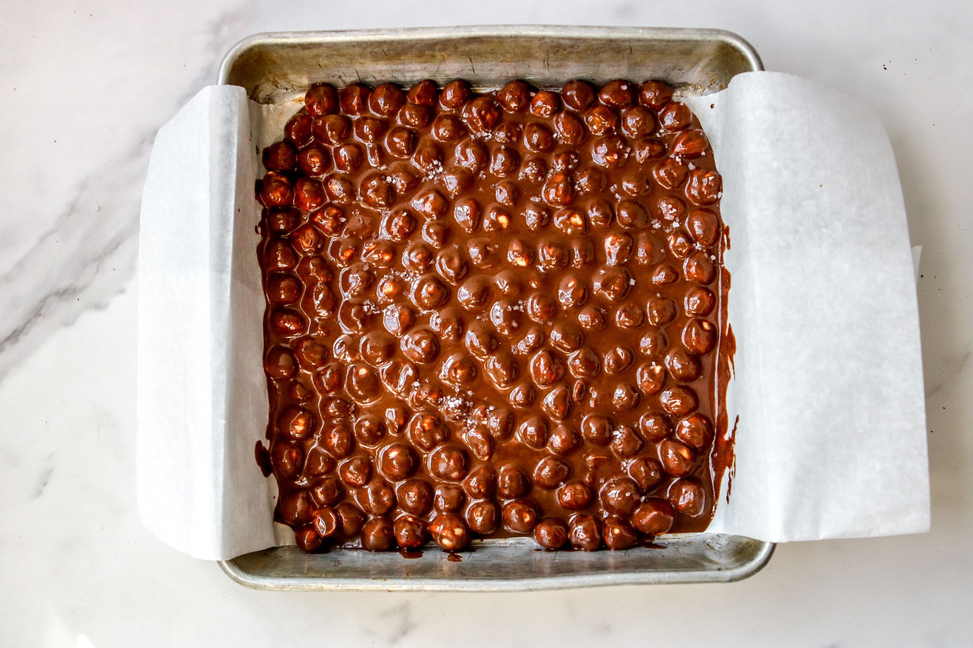 This is an overhead horizontal image of a square pan lined with white parchment paper. In the pan, on top of the parchment paper is a layer of hazelnuts coated in chocolate across the bottom of the pan. The pan sits on a white marble counter.