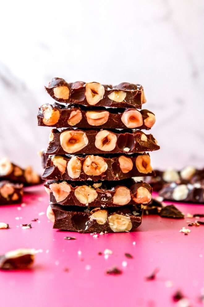 This is a vertical image looking at a stack of chocolate bars from the side. The chocolate has tons of hazelnuts in it. The stack sits on a dark pink counter with more chocolate pieces and crumbs blurred in around the stack and in the background. The stack sits against a white background. 