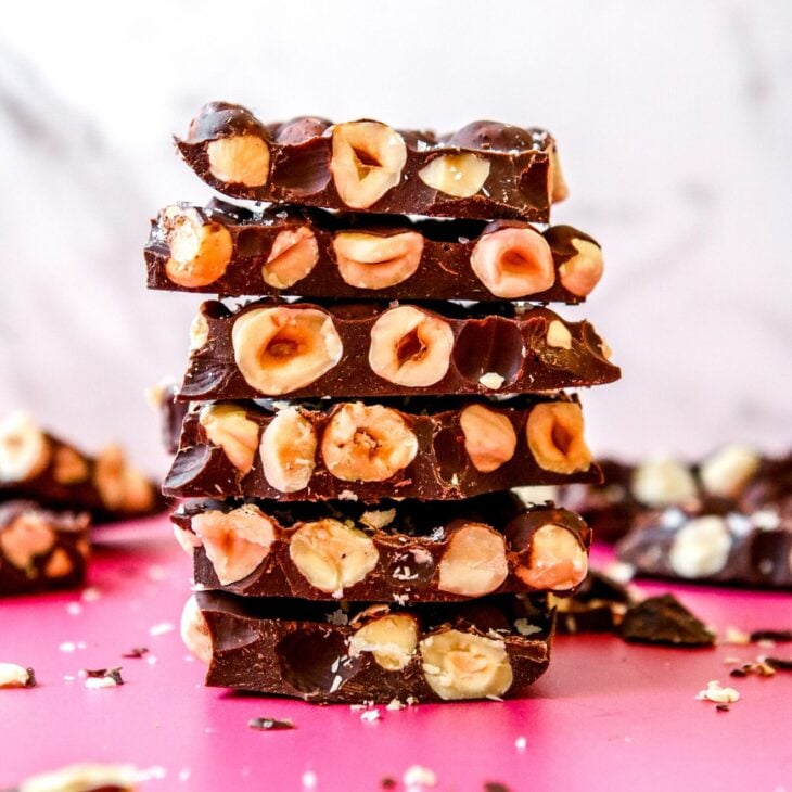This is a vertical image looking at a stack of chocolate bars from the side. The chocolate has tons of hazelnuts in it. The stack sits on a dark pink counter with more chocolate pieces and crumbs blurred in around the stack and in the background. The stack sits against a white background.