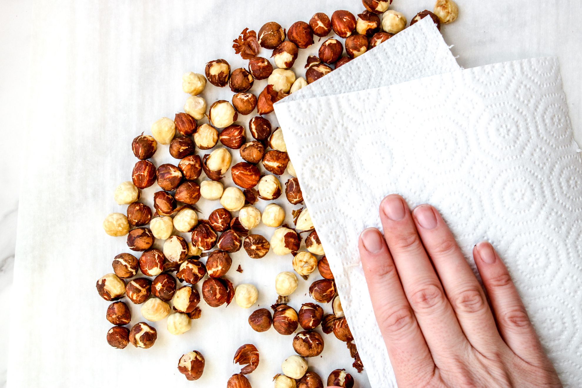 This is an overhead horizontal image of roasted hazelnuts on a white pieces of parchment paper. A hand is holding a paper towel on top of the hazelnuts to rub the skins off. 