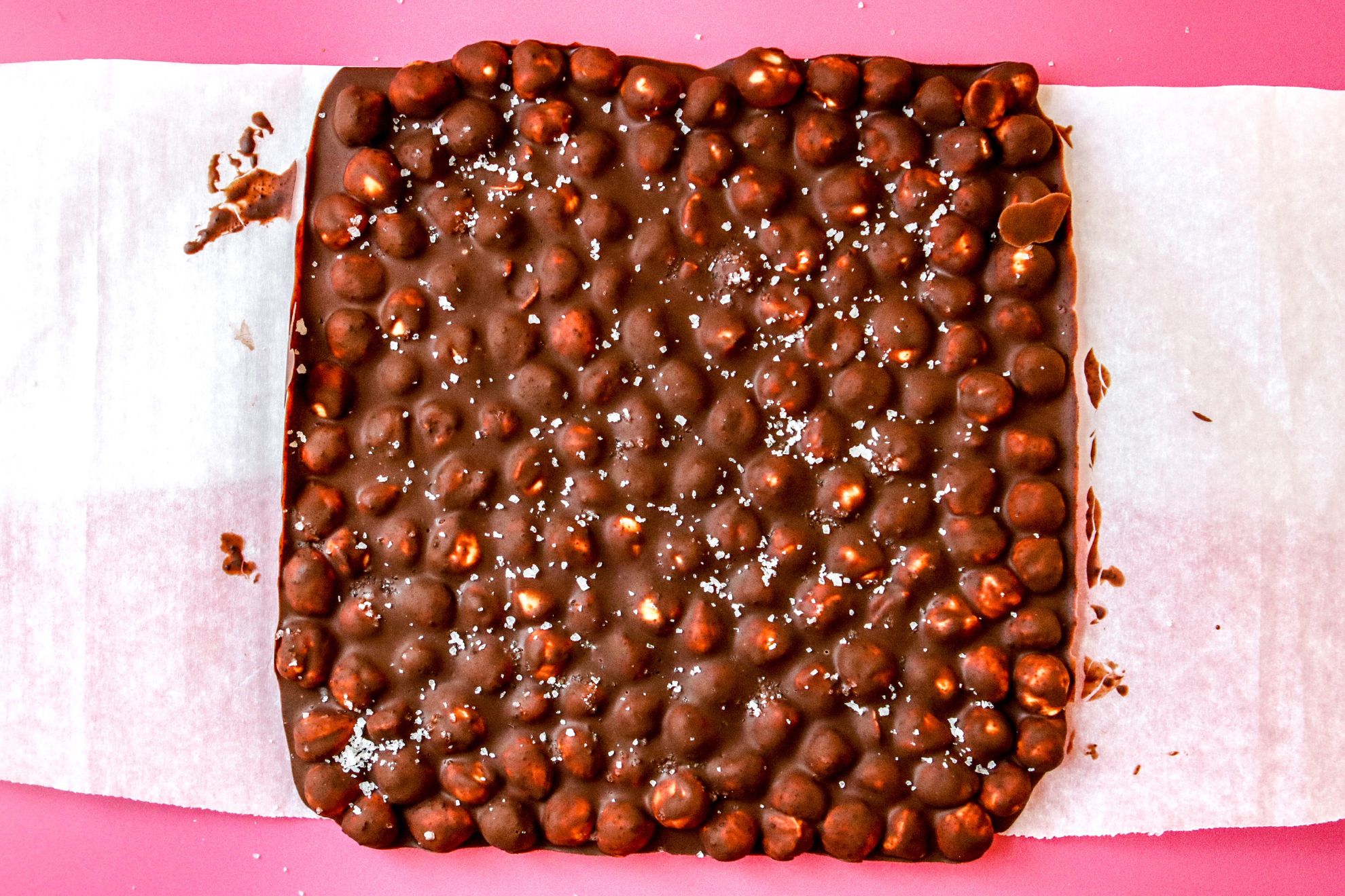 This is an overhead horizontal image of a square of chocolate with hazelnuts in it and flakey salt on top. The square oif chocolate sits on a white piece of parchment paper on top of a dark pink surface.
