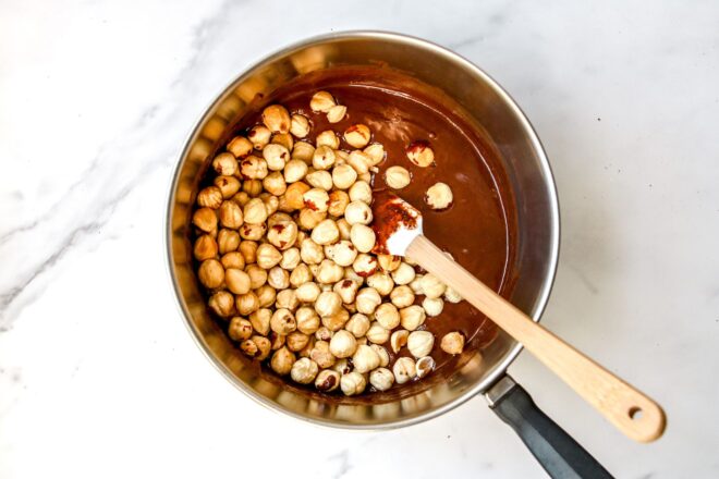 This is an overhead image of a saucepan with melted chocolate and hazelnuts in it. A small white spatula is dipped into the chocolate with the wooden handle leaning against the side to the bottom right corner of the image. The pot sits on a white marble surface. 