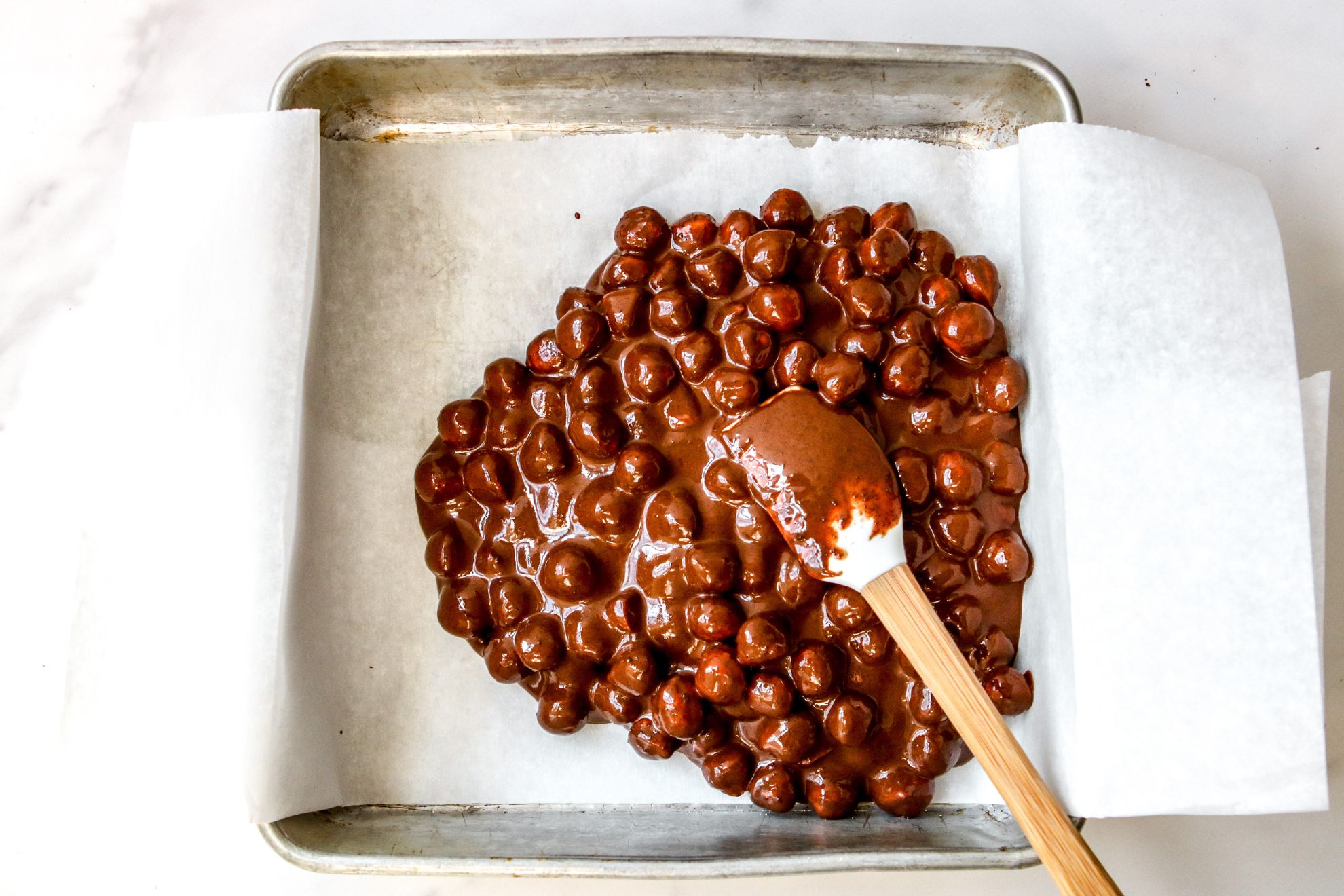 This is an overhead horizontal image of a square pan lined with white parchment paper. In the pan, on top of the parchment paper are hazelnuts coated in chocolate in the center and to the right of the pan. A small white spatula is coated in chocolate and lying on top of the mixture with the wooden handle leaning against the side of the pan, to the bottom right corner of the image. The pan sits on a white marble counter.