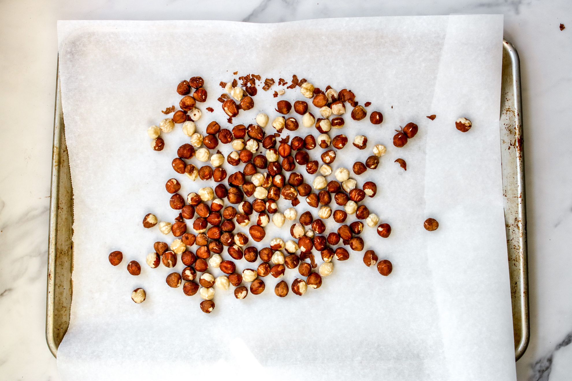 This is an overhead horizontal image of a baking sheet lined with parchment paper. On top of the parchment paper are hazelnuts with their skins on, spread out in an even layer. The baking sheet sits on a white marble counter. 