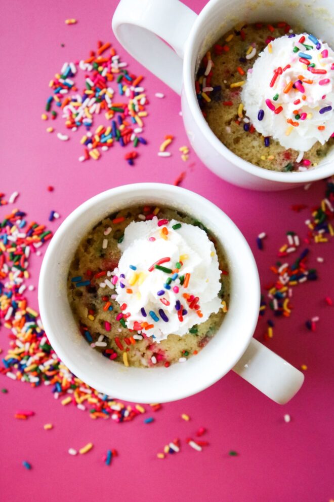 This is an overhead vertical image of two white mugs with funfetti sprinkle cake in it. The mug cake has rainbow sprinkles mixed throughout the cake, is topped with whipped cream with more rainbow sprinkles sprinkled on top. The image focuses on one white mug in the center with the second mug cut off in the bottom right corner of the image. The white mugs sit on a dark pink surface with rainbow sprinkles scattered around them.