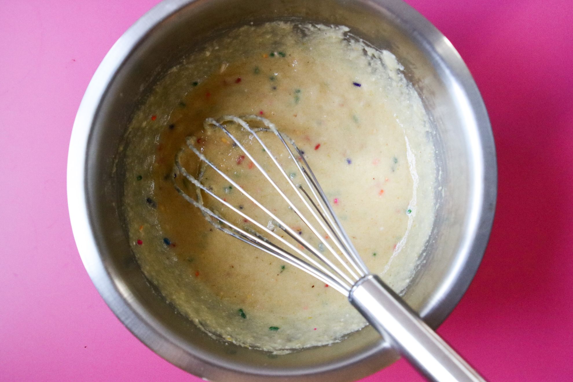 This is an overhead horizontal image of a stainless steel bowl with a vanilla batter with rainbow sprinkles mixed in. A whisk is in the batter with the handle of the whisk leaning against the side of the bowl and angled to the bottom right corner of the image. The bowl sits on a dark pink surface.