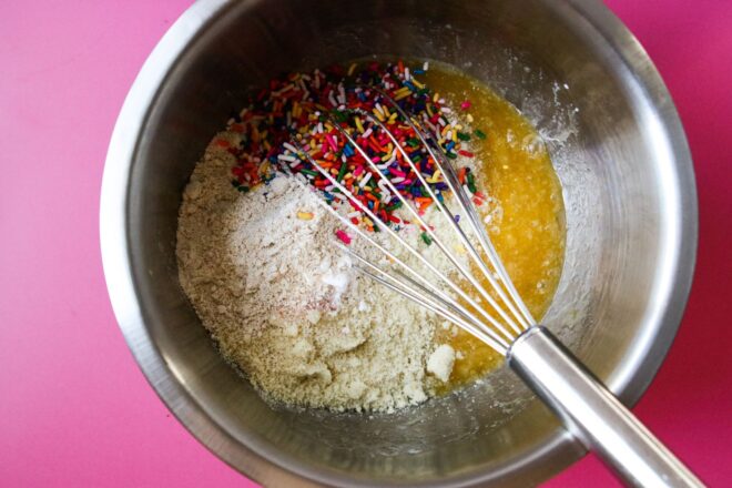 This is an overhead horizontal image of a stainless steel bowl with wet ingredients mixed together and dry ingredients on top, not yet mixed in. There appears to be two different kinds of dry ingredients and rainbow sprinkles. A whisk is in the bowl and leaning against the side with the handle to the bottom right corner of the image. The bowl sits on a dark pink surface.