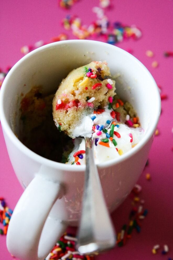 This is a vertical image peering into a white mug with vanilla sprinkle cake in it. The sprinkle cake is topped with whipped cream and more rainbow sprinkles. A spoon is digging into the cake and leaning against the side of the mug. The scoop of the spoon sits on top of the cake and has a bite of fluffy vanilla sprinkle cake in it. The mug sits on a dark pink surface with rainbow sprinkles scattered around it.