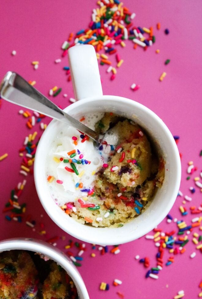 This is an overhead vertical image of two white mugs with funfetti sprinkle cake in them. The mug cake has rainbow sprinkles mixed throughout the cake, is topped with whipped cream with more rainbow sprinkles. The image focuses on one white mug in the center with the second mug cut off in the bottom left corner of the image. Silver spoons are digging into the cake and leaning against the side of the mug. The white mugs sit on a dark pink surface with rainbow sprinkles scattered around them.