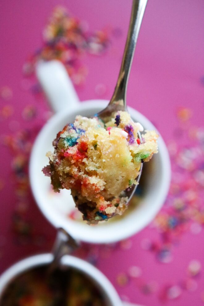 This is an overhead vertical image of a silver spoon holding a bite of funfetti cake. Blurred behind the spoon is one white mug in the center of the image and another mug to the bottom left corner of the image. The mugs sit on a dark pink surface with rainbow sprinkles scattered around them.