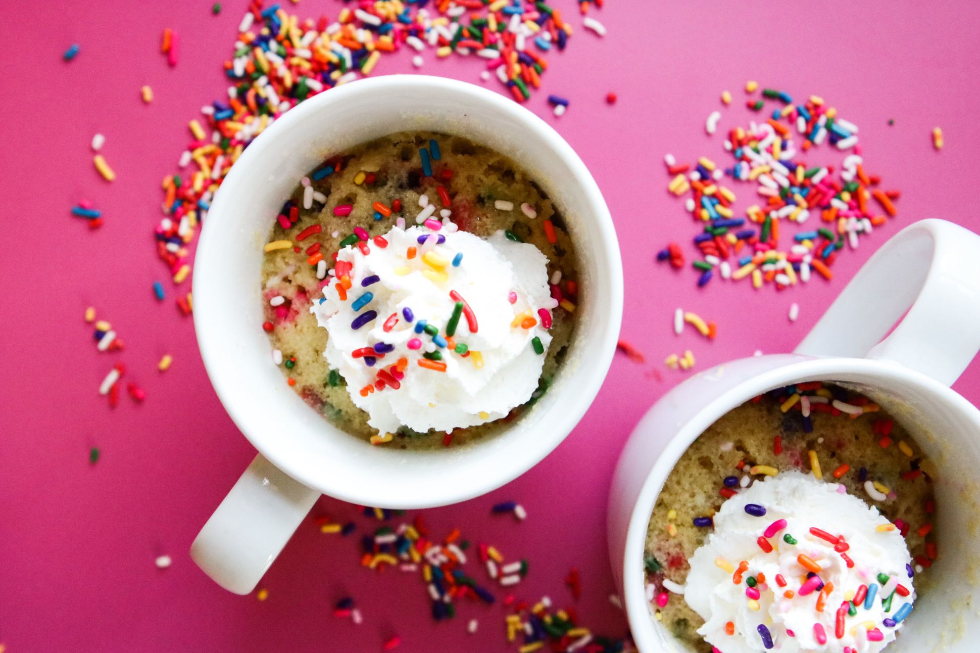 This is an overhead horizontal image of two white mugs with funfetti sprinkle cake in it. The mug cake has rainbow sprinkles mixed throughout the cake, is topped with whipped cream with more rainbow sprinkles sprinkled on top. The image focuses on one white mug in the center with the second mug cut off in the bottom right corner of the image. The white mugs sit on a dark pink surface with rainbow sprinkles scattered around them.