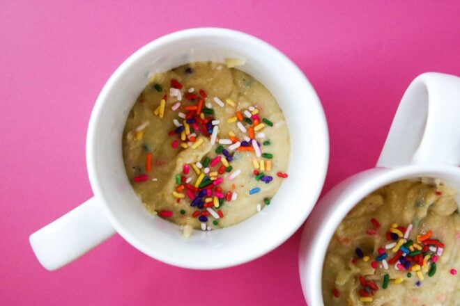 This is an overhead horizontal image of two white mugs with raw vanilla cake batter in it. The batter has rainbow sprinkles mixed in and more sprinkled on top. The image focuses on one mug with a second mug cut off in the bottom right corner of the image. The white mugs sit on a dark pink surface.