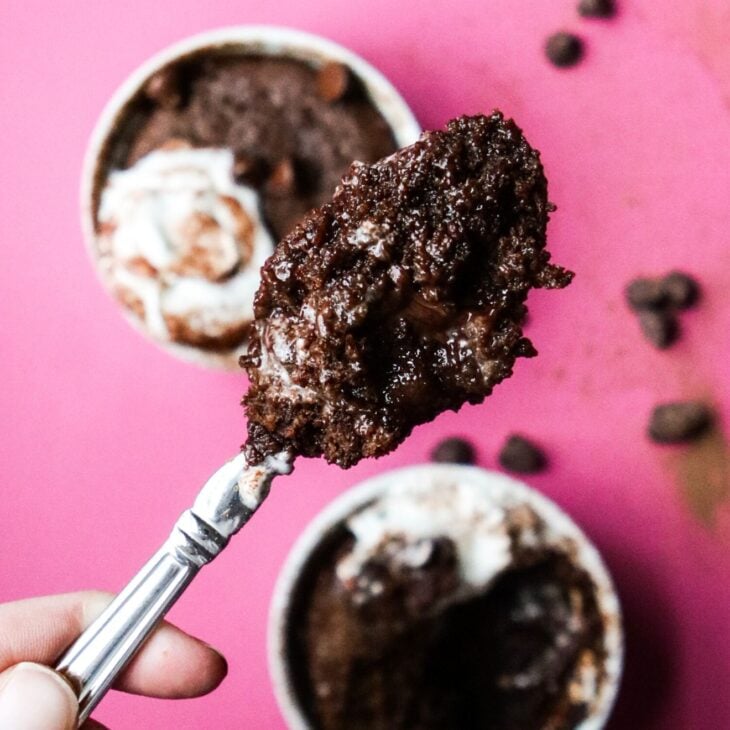 This is an overhead vertical image of a hand holding a silver spoon with gooey chocolate cake on it. Blurred in the background are two white ramekins sitting on a dark pink surface. In the ramekins are chocolate cake topped with whipped cream and chocolate chips. More chocolate chips and some cocoa powder are scattered around the white ramekins.
