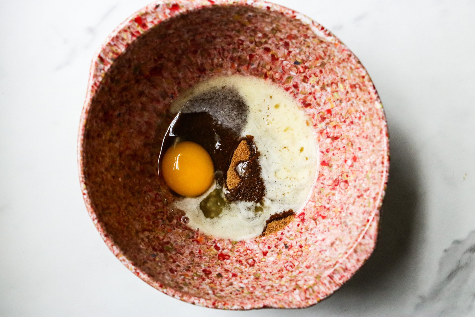 This is an overhead horizontal image of a pink speckled bowl on a white marble counter. In the bowl are wet ingredients including butter, coconut sugar, and an egg.