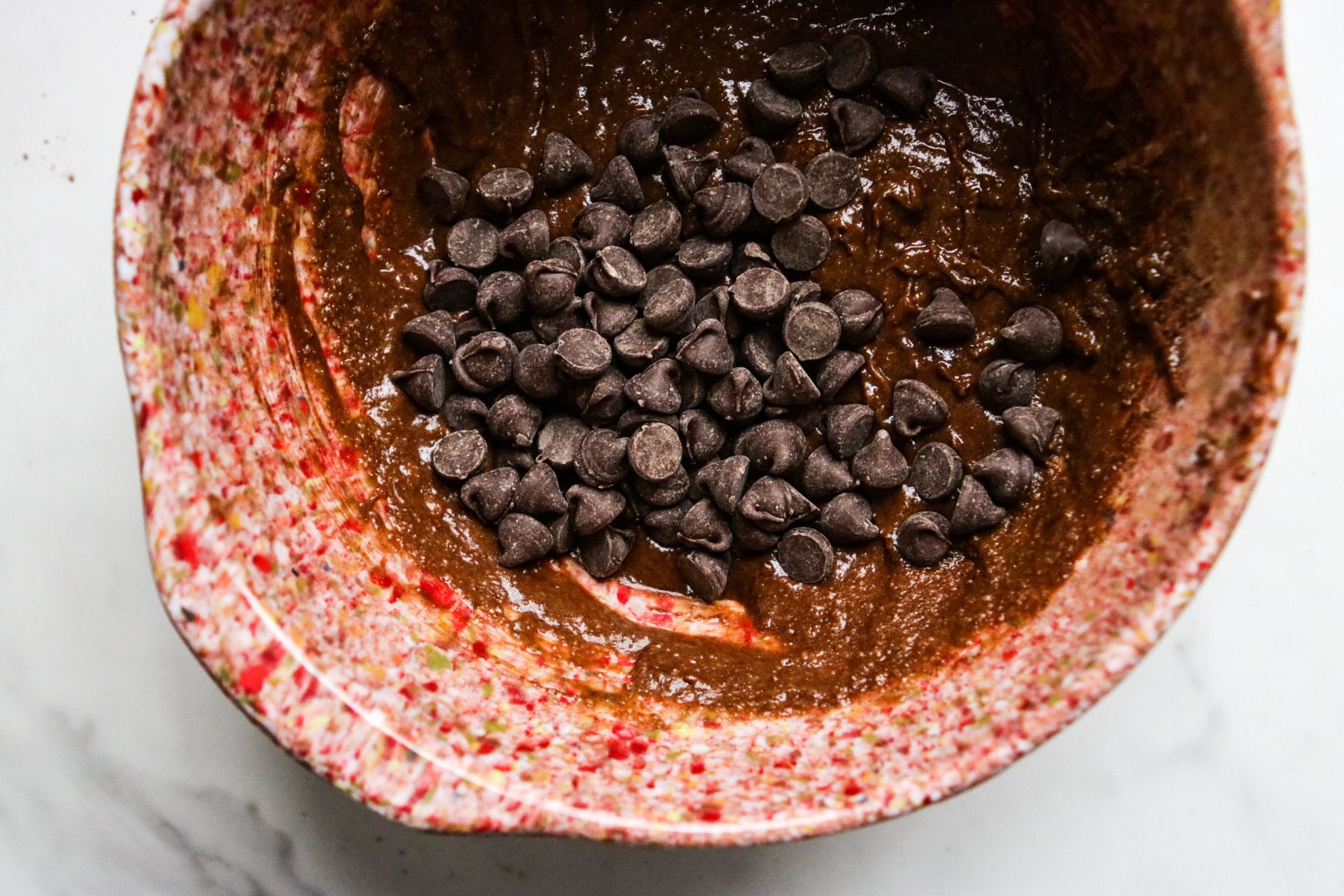 This is an overhead horizontal image of a pink speckled bowl on a white marble counter. In the bowl is a chocolate batter with chocolate chips on top.