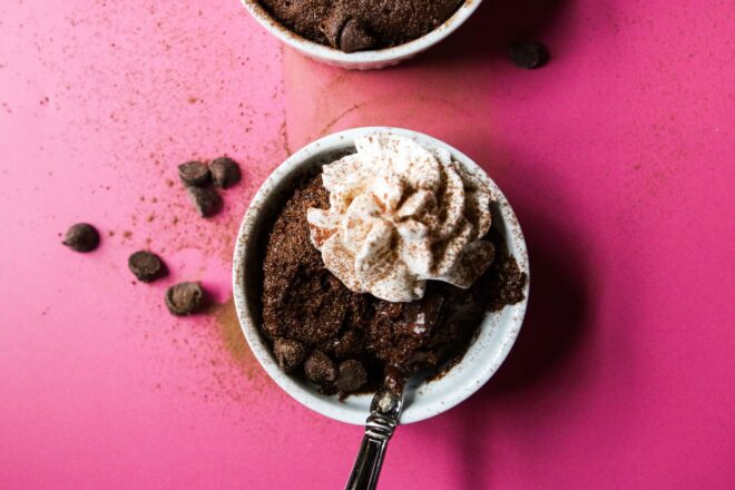This is an overhead horizontal image of a white ramekin with chocolate cake in it. The cake is topped with a dollop of whipped cream and cocoa powder. A spoon is digging in and leaning against the side of the ramekin with the handle pointing toward the bottom. The ramekin is on a deep pink surface with chocolate chips and cocoa powder scattered around it. Another ramekin is coming from the top of the image.