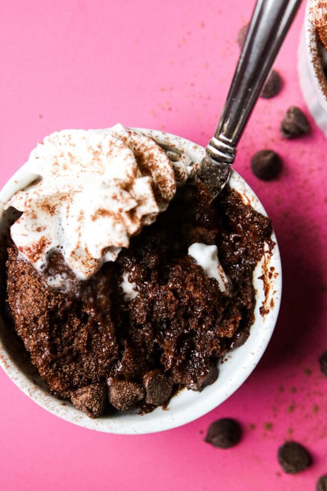 This is an overhead vertical image of a white ramekin with chocolate cake in it. The cake is topped with a dollop of whipped cream and cocoa powder. A spoon is digging into the gooey cake and leaning against the side of the ramekin with the handle pointing toward the top right corner. The ramekin is on a deep pink surface with chocolate chips and cocoa powder scattered around it. Another ramekin is coming from the top right corner of the image.