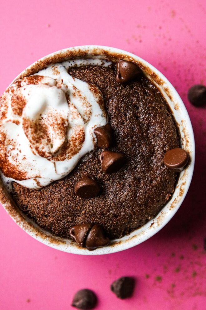 This is a vertical overhead image of a white ramekin with chocolate cake in it. The cake is topped with a dollop of whipped cream, cocoa powder, and chocolate chips. The ramekin is on a deep pink surface with chocolate chips and cocoa powder scattered around it.