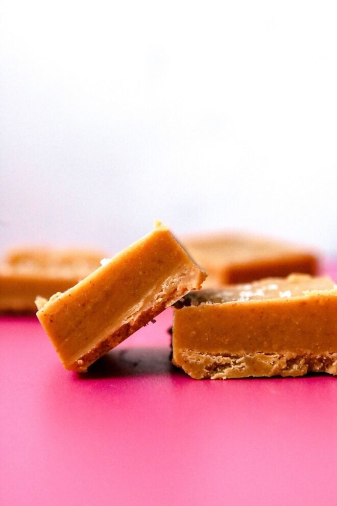 This is a vertical image looking at two peanut butter fudge squares from the side. One square is leaning against another square to the right. The pieces of fudge sit on a deep pink surface with more peanut butter fudge pieces blurred in the white background.