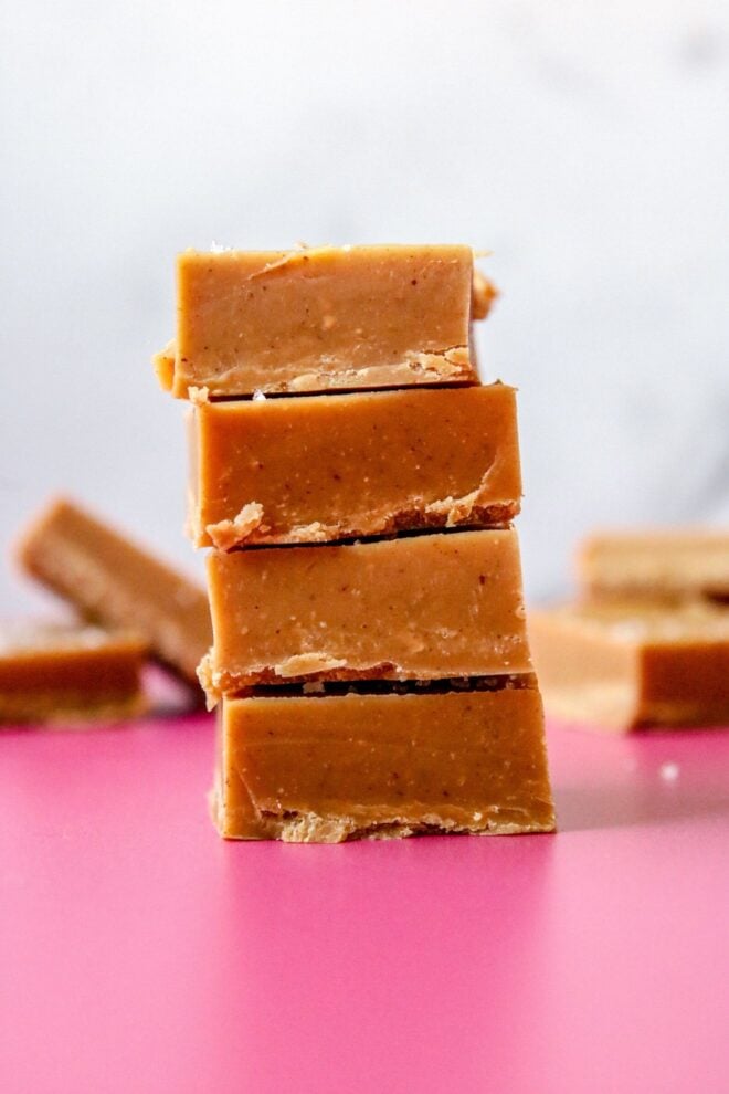 This is a vertical image looking from the side at a stack of 4 peanut butter fudge squares stacked. The stack sits on a deep pink surface with more squares blurred in the white background.