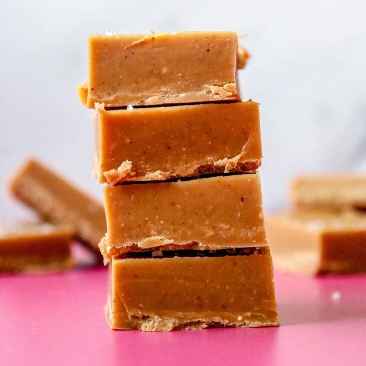This is a vertical image looking from the side at a stack of 4 peanut butter fudge squares stacked. The stack sits on a deep pink surface with more squares blurred in the white background.