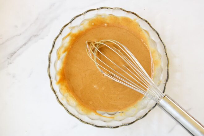 This is an overhead horizontal image of a glass bowl with a peanut butter mixture in it. A whisk is in the bowl and leaning against the side of the bowl angled to the bottom right corner of the image. The bowl sits on a white marble counter.