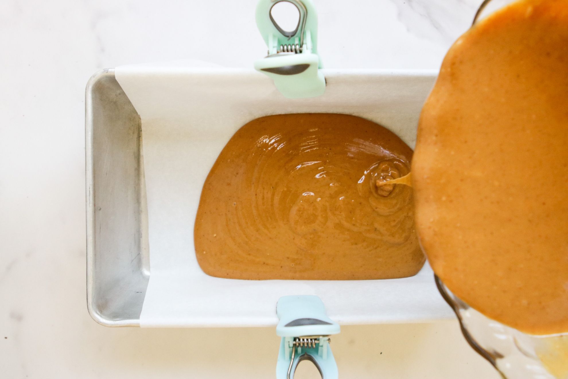 This is an overhead horizontal image of a bread pan lined with parchment paper secured with light blue and green chip clips on the side. The pan sits on a white marble counter. A peanut butter mixture is being poured from a glass bowl coming from the right side of the image. The peanut butter mixture is being poured into the pan and a puddle of peanut butter mixture is in the bottom of the pan.