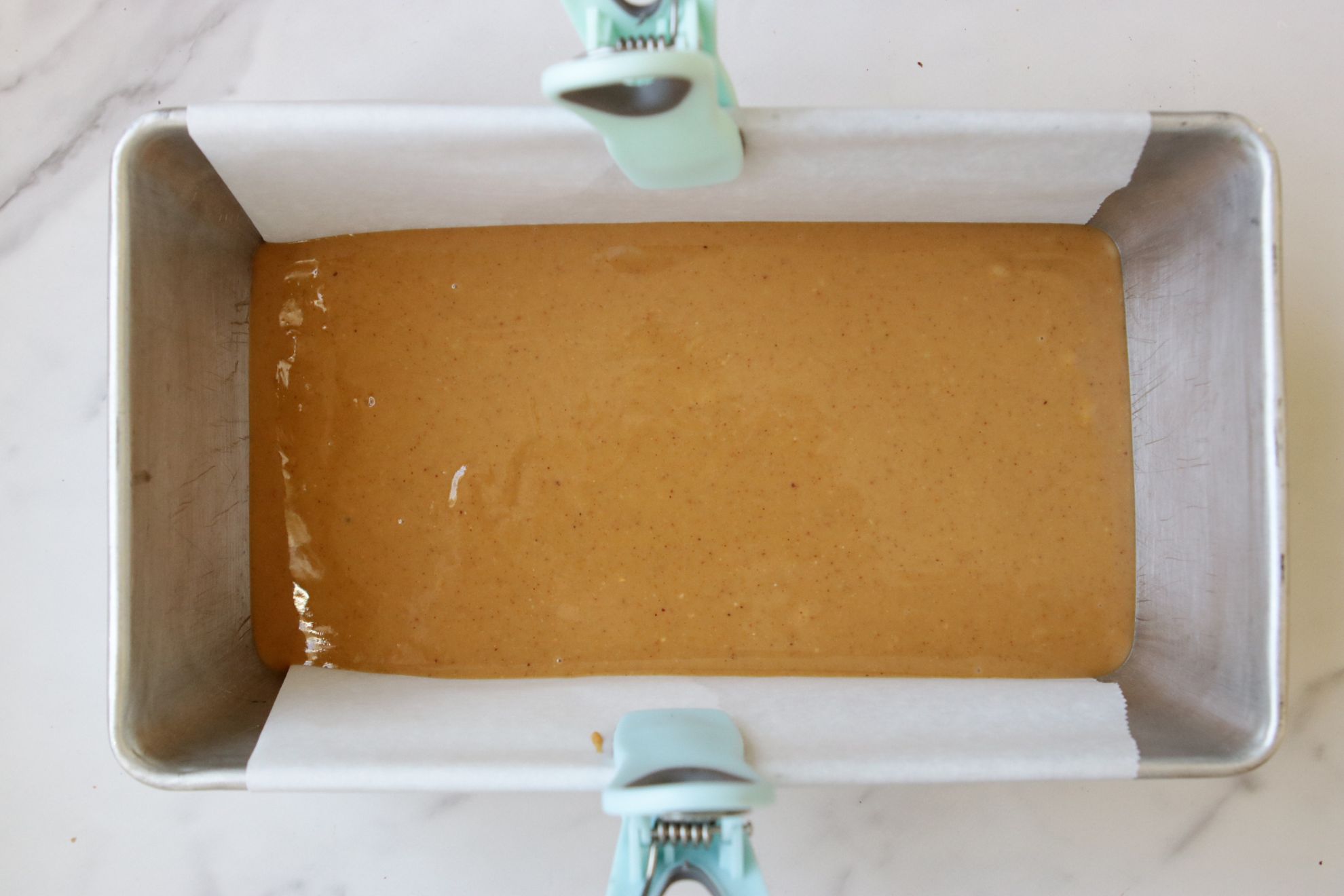 This is an overhead horizontal image of a bread pan lined with parchment paper and secured on the long side of the pan with light blue and green plastic chip clips. In the pan is a melty peanut butter mixture. The pan sits on a white marble counter.