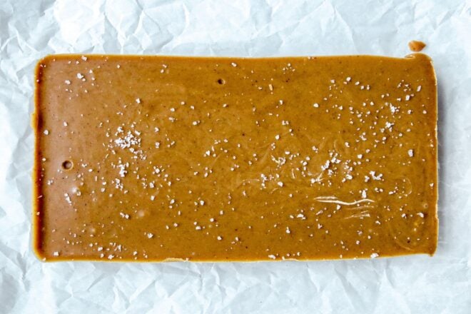 This is an overhead horizontal image of a rectangle of hardened peanut butter sprinkled with salt. The peanut butter fudge sits on a crinkled white piece of parchment paper.