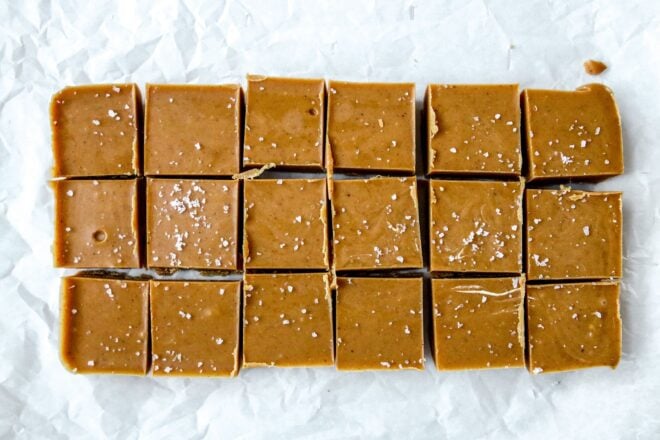 This is an overhead horizontal image of a rectangle of hardened peanut butter fudge sprinkled with salt. The peanut butter fudge is cut into 18 squares and sits on a crinkled white piece of parchment paper.