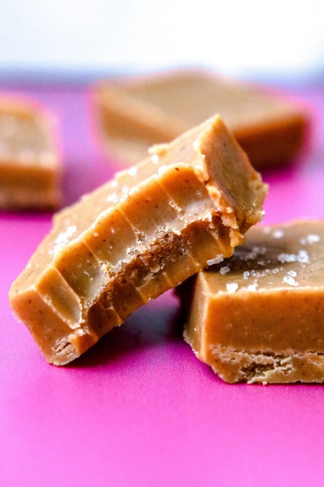 This is a vertical image of a side view looking at a square of peanut butter fudge with a bite taken out of it. The fudge square leans against another square to the right of the image. The fudge squares sit on a hot pink surface with more fudge blurred in the background.