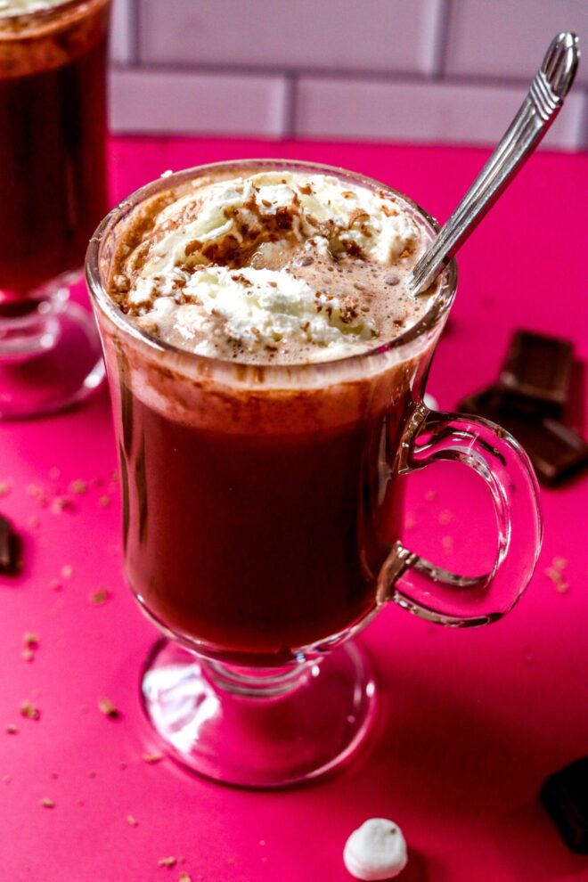 This is an overhead image shot from an angle of hot chocolate with whipped cream in a glass mug. A silver utensil is in the hot chocolate and leaning against the side of the mug. The mug sits on a hot pink surface with pieces of a broken chocolate bar on the pink surface surrounding the mug. Another bottom of a glass mug is blurred in the background with a few mini marshmallows to the top right corner of the image.