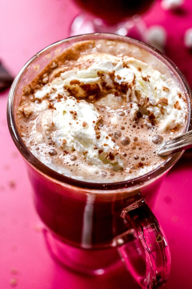 This is an overhead image shot from an angle of hot chocolate with whipped cream in a glass mug. A silver utensil is in the hot chocolate and leaning against the side of the mug. The mug sits on a hot pink surface with pieces of a broken chocolate bar on the pink surface surrounding the mug. Another bottom of a glass mug is blurred in the background with a few mini marshmallows to the top right corner of the image.