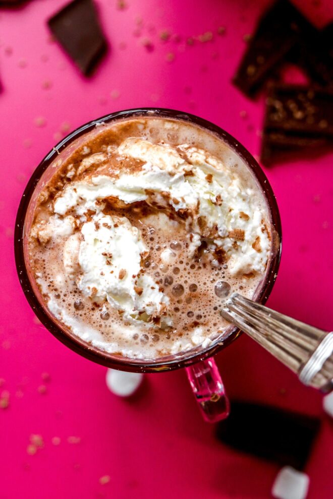 This is an overhead image of hot chocolate with whipped cream in a glass mug. A silver utensil is in the hot chocolate and leaning against the side of the mug. The mug sits on a hot pink surface with pieces of a broken chocolate bar on the pink surface surrounding the mug.