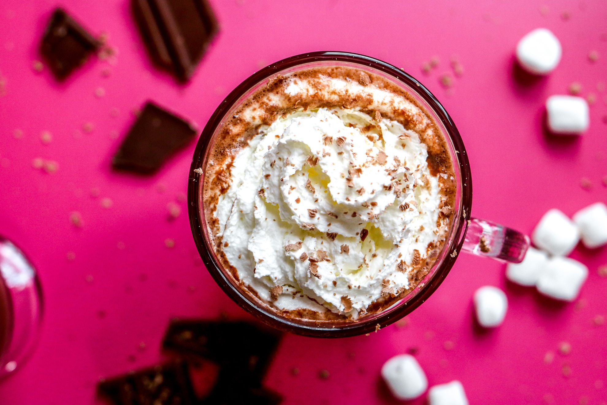 This is an overhead image of hot chocolate with whipped cream in a glass mug. The mug sits on a hot pink surface with pieces of a broken chocolate bar and mini marshmallows on the pink surface surrounding the mug.