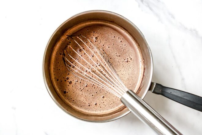 This is an overhead image of saucepan with hot chocolate in it and a white. The saucepan sits on a white marble surface.